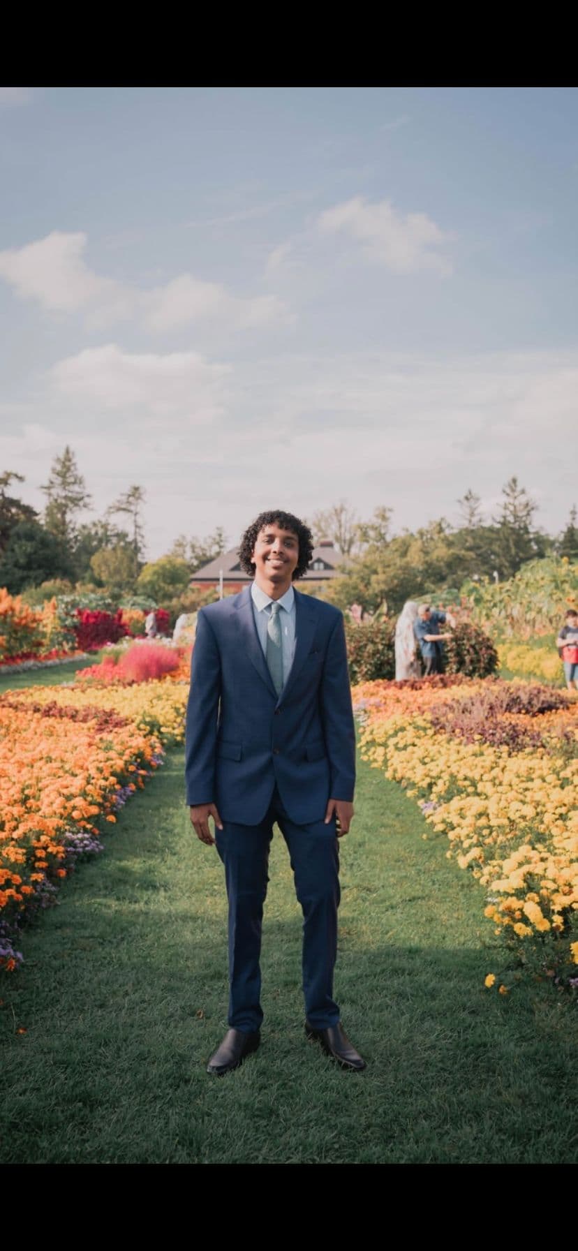 Smiling man in a blue suit standing on a grass path between vibrant flower beds.